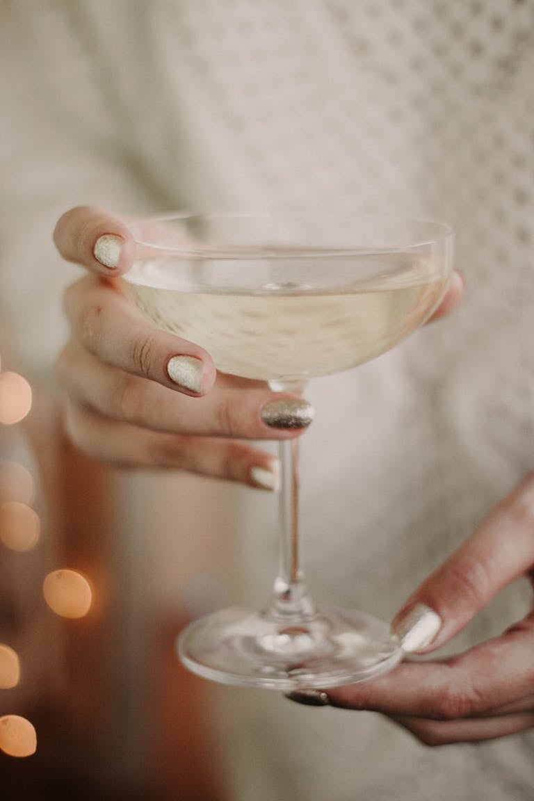 Crop unrecognizable female with shiny nails in white sweater enjoying champagne from elegant glass